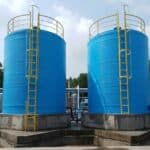 Dua unit tangki fiberglass besar berwarna biru dengan tangga akses kuning di area pabrik industri. Terdapat teks 'TANGKI FIBERGLASS', 'AWET KOKOH TAHAN LAMA', dan logo 'TOYA ARTA SEJAHTERA'. Menunjukkan produk tangki penyimpanan berkualitas tinggi untuk kebutuhan industri.