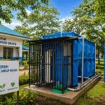 Pemandangan luar fasilitas Dapur Makan Bergizi Gratis dengan instalasi pengolahan air bersih tangki biru modern dan papan tanda bertuliskan 'Air Bersih Untuk Hidup Sehat' di bagian depan.